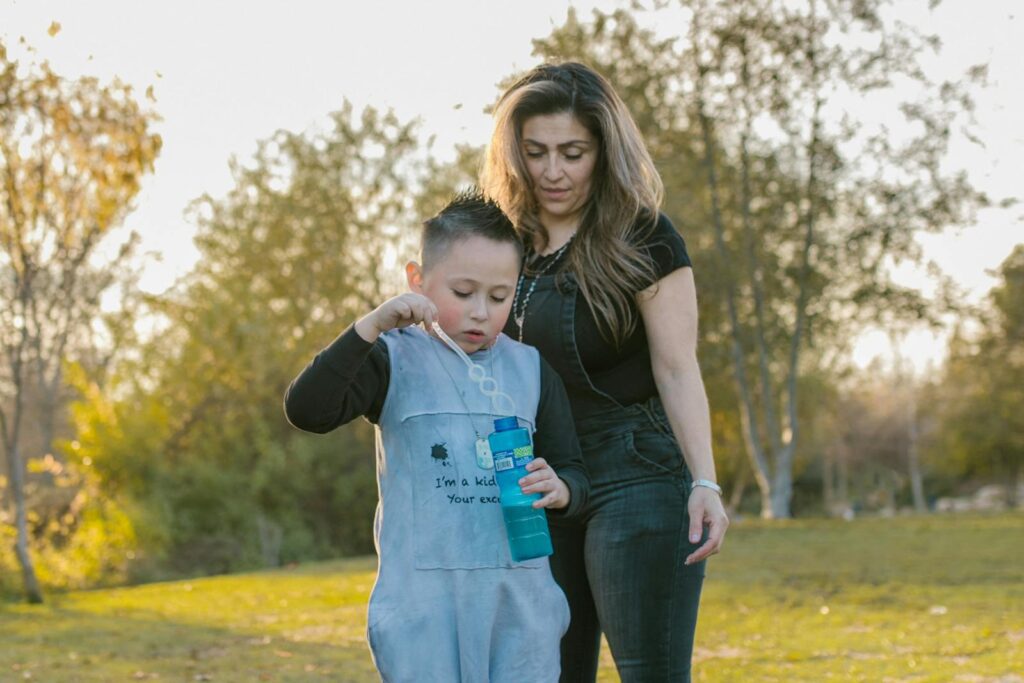 A mother and son enjoy blowing bubbles together in a sunny park setting.