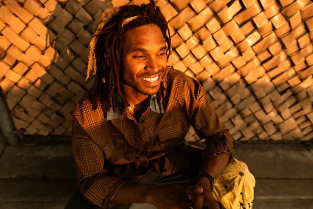 Smiling man with dreadlocks enjoying the sunset outdoors, exuding warmth and happiness.